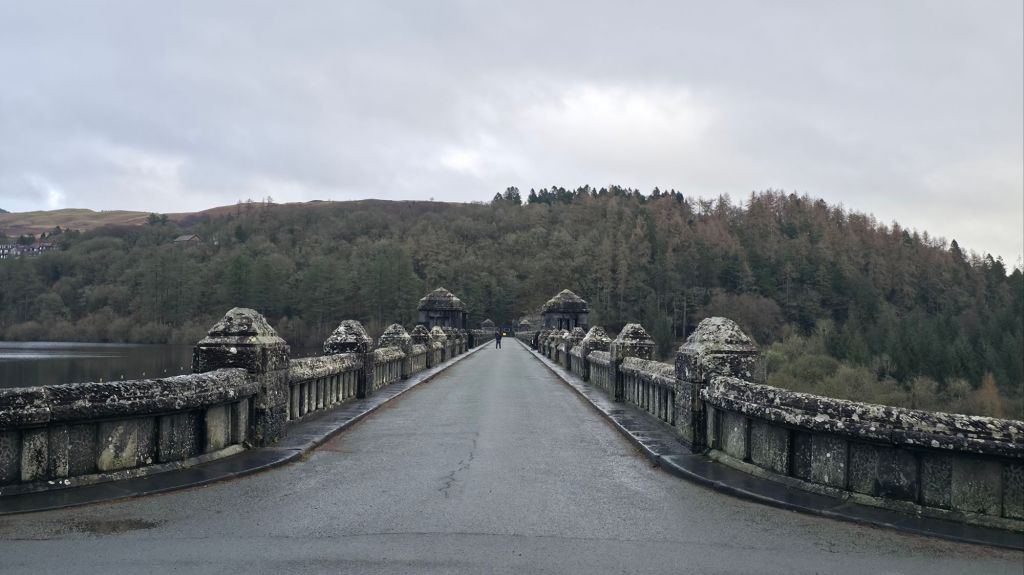 Widok na kamienny most prowadzący przez zbiornik wodny, w tle zalesione wzgórza i szare niebo.